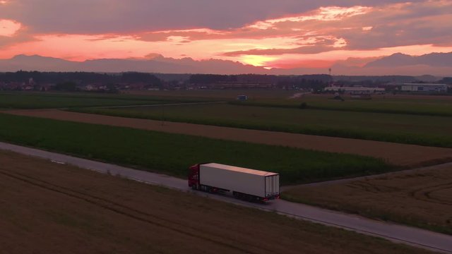 AERIAL: Flying along a cargo truck transporting goods across the picturesque countryside at golden sunrise. Lorry drives goods towards a logistics center in the countryside on a sunny summer evening.