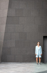 Full-length portrait of young stylish woman standing near modern building and looking at camera. Concept of fear and loneliness. Vertical shot