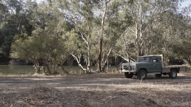 A pick up truck drives slowly through frame along a dirt road next to a tree lined river in country Victoria. The camera tracks with the truck from right to left.