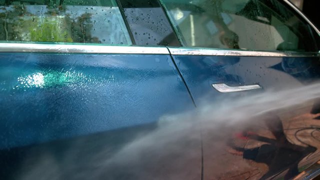 SLOW MOTION, CLOSE UP: Water is sprayed over side of a blue car being washed in the driveway on sunny summer afternoon. Jet washer squirts water over the shiny car door. Detailed shot of washing a car