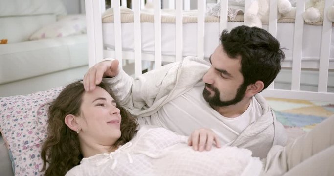 Man and Pregnant Woman Sitting on Floor, Embracing and Laughing. Young Family Expecting a Baby. White Interior with Baby Cot on the background. Concept of Parenthood, Happiness and Tenderness