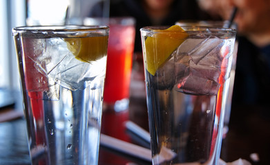 Glasses of refreshing ice cold water with lemons on restaurant table with patrons in the background.