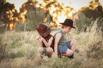 Young boys dressed up as cowboys in an open field with long grass and a vibrant sunset in the...