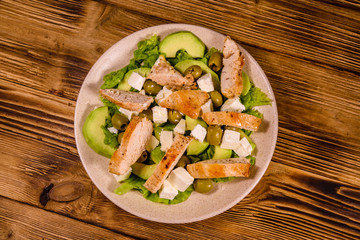 Fresh salad with chicken meat, feta cheese, avocado, green olives and lettuce leaves in ceramic plate on wooden table. Top view