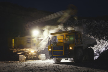 Excavator loads a large mining dump truck on a dark background in the night shift, close-up.