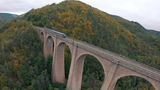Follow, wide angle aerial view of a French SNCF train crossing an old stone bridge in a beautiful natural valley with colorful trees in  an autumn landscape. Shot with an FPV drone.