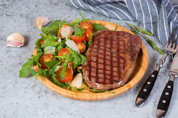 Close up Grilled beef steak with fresh arugula, tomato and Parmesan salad on a plate on a grey concrete background. Horizontal photo