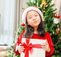 Little asian girl smile and excited and holding red gift box on christmas tree and white...