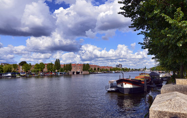 Fototapeta premium Amsterdam, Holland, August 2019. View of the Amstel River, outskirts of the city. A large moored boat is used as a houseboat. Sunny day with blue sky and white clouds.