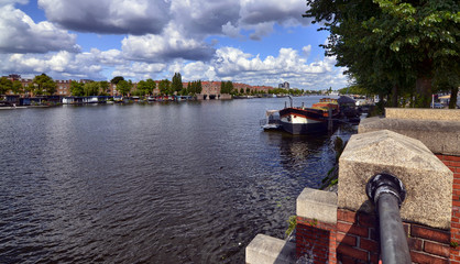 Naklejka premium Amsterdam, Holland, August 2019. View of the Amstel River, outskirts of the city. A large moored boat is used as a houseboat. Sunny day with blue sky and white clouds.