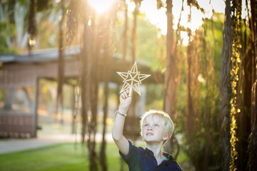 Young boy holding gold Christmas star up in the air with natural sunlight streaming through