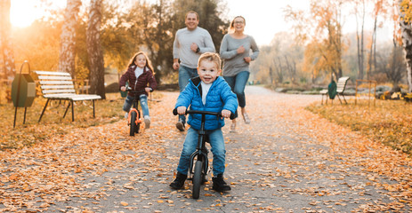 Obraz premium Lovely little kid running away on his byclies while her parents and sister is trying to catch him outdoor in the park.