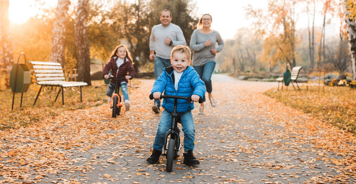 Lovely Little Kid Running Away On His Byclies While Her Parents And Sister Is Trying To Catch Him Outdoor In The Park.