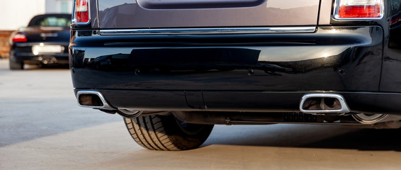Close-up of a black luxury car bumper of an sedan with two side turbo exhaust pipes outdoors on asphalt. Auto service industry. Air and environment pollution.