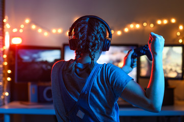 Gamer or streamer girl at home in a dark room with a gamepad playing video games with friends online. Young man sits in front of a monitor - obrazy, fototapety, plakaty