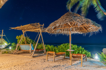 Wooden swing with a mattress under a canopy and straw umbrella on the tropical beach near sea at night, island Zanzibar, Tanzania, Africa