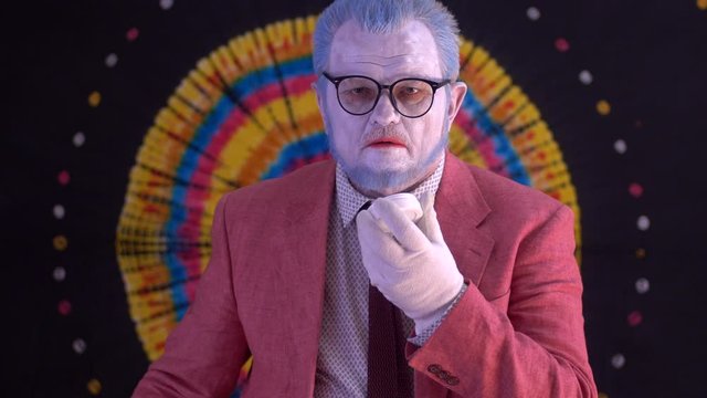 elderly mime man in large glasses with blue hair and beard in bright pink jacket and tie in white gloves. clown does makeup on face. White face, blue hair, red lips. Happy clown