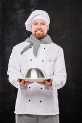 Young smiling chef in uniform holding cloche with cooked meal for you