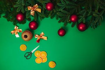 Christmas hand made how to make Christmas wreath. Scissors, rope, red christmas balls, golden bows with fir branches and dried oranges on the green background.