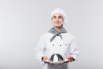 Happy young successful female chef in uniform carrying cloche with cooked meal
