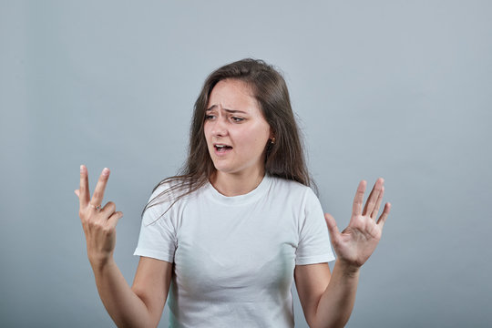 Girl in white t-shirt looks at her three fingers, with another hand shows woman five. Behind her there is gray background. Teen looks cofused.