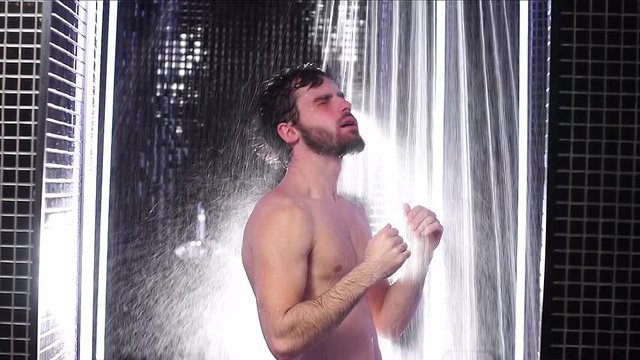 Young caucasian bearded male taking shower indoors. Fit muscular man enjoy and relax in shower. Slow motion