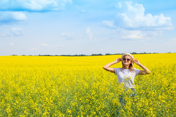 Smiling girl in sunglasses, holds a hat with her hands and stands in a bright yellow field