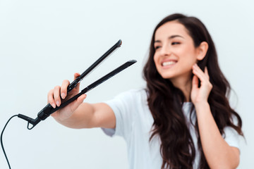 Pretty, smiling girl holding a straightener in her hands. Closeup, blurred background