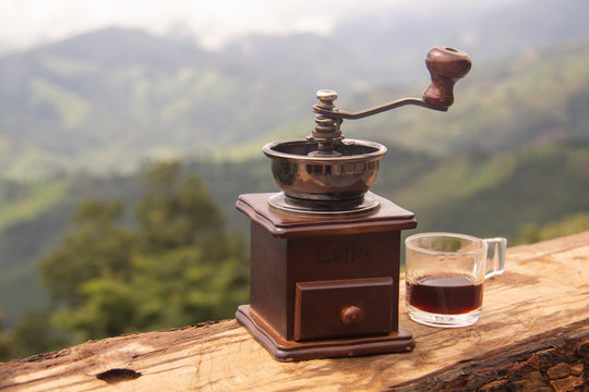 An ancient coffee grinder placed on a wooden table with a mountain backdrop and a cup of coffee next to it.