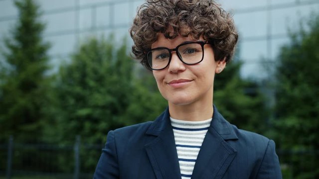 Smiling ambitious girl in suit and glasses standing in city center near office building looking at camera with cheerful face. Confidence and modern people concept.