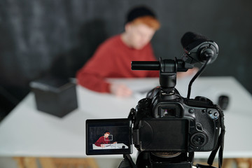 Screen of video camera in front of young male vlogger sitting by desk