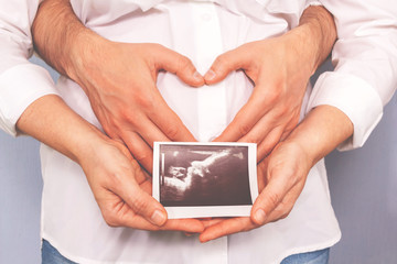 Young couple making heart shape on the pregnant belly. Minimalism lifestyle. Woman holding photo ultrasound. Amazing, sweet, toned photo. Pregnancy, expecting a baby concept.