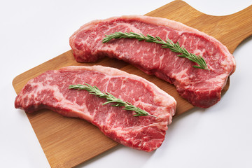 Raw juicy beef steak ready to be cooked. Close up photo of two fresh raw striploin steak on wooden board on white background with rosmary. Top view.