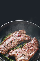 Steaming, reeky, flavored spicy grilled beef striploin steak in a frying pan on a black background with copy space. Frying pan, steak, cooking on fire concept
