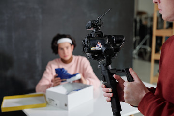 Young man holding video camera while standing in front of vlogger by desk