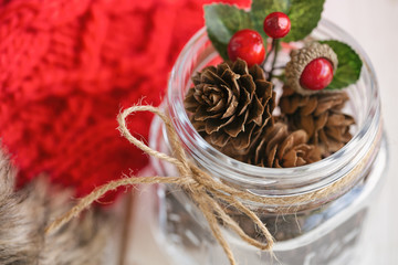 Pine cone or conifer cone and red holly balls in glass bottle with rustic ribbon bow tie. Christmas background with festive decoration red knitwear wool gloves for Christmas wallpaper.