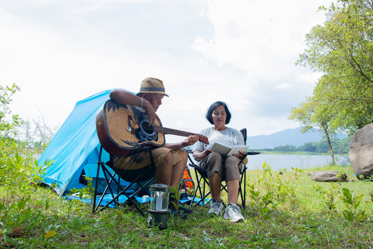 An elderly couple in Asian comes to travel and camping to relax with guitar on a holiday near a stream that looking beautiful natural alternating. Happiness of lover concept.