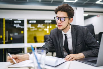 Young office manager working with papers and computer working at office. Businessman writes, reviews and analyze documents, plans. Stylish man in a suit working with new startup project.