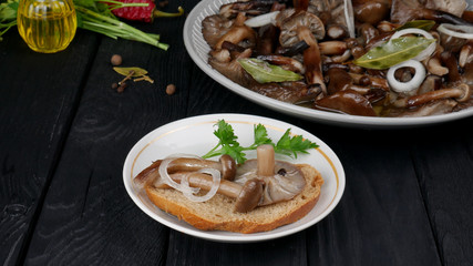 Sandwich with pickled mushrooms, a ring of onion and leaf of parsley. Pickled forest mushrooms in a plate on a black wooden table with butter and spices in the background