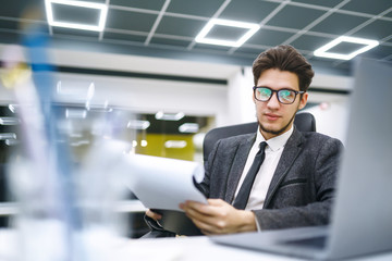 Fototapeta premium Young office worker in glasses reads documents in the modern office. Young business manager working with new startup project. Stylish man in a suit reviews and analyze documents, plans.