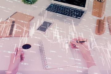 Hands writing information about stock market in notepad. Forex chart holograms in front. Concept of...