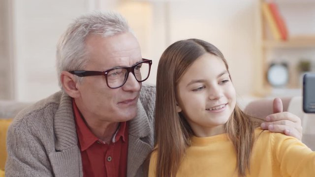 Chest-up shot of senior Caucasian granddad with grey hair, in glasses, and young granddaughter sitting together on couch at home with smartphone, both looking at it, hugging, smiling and talking