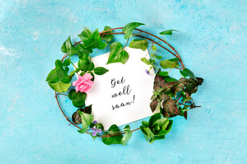 Get well soon card with a wreath of green ivy leaves and a tender pink rose, overhead shot on a blue background
