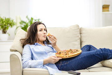 Tired woman is chewing pizza, while laying on the white sofa. She is watching TV shows, being on...