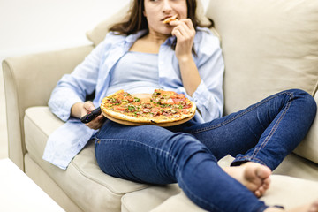 Crop. Close up legs in jeans. Pretty woman is laying on the sofa at home and is watching television while eating pizza.