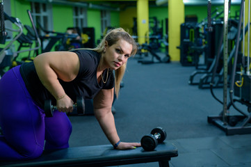 An obese woman is training with dumbbells on a bench in the gym. The fat blonde is losing weight with the help of fitness.