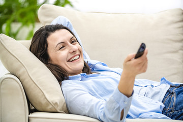Happy young woman is watching tv, sitting on a sofa. She is smiling widely. Crop.