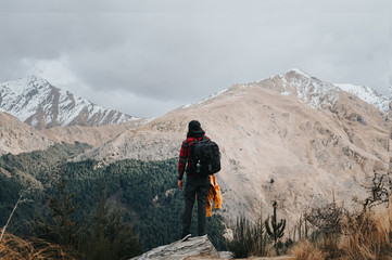 Man hiking on the peak of mountain with pine forest and snow capped mountains in background.