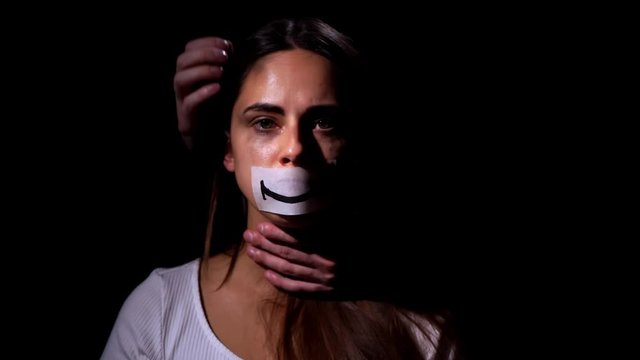 Domestic violence and social problems concept. Desperate woman in depression looks at the camera and puts a smile image on her face on black background
