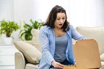 Brunette woman is opening the pizza box, isolated, sitting on the light couch.